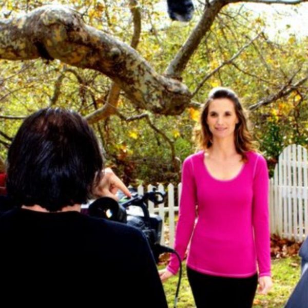 Karena Thek being filmed outdoors by a camera crew in a garden setting with autumn foliage and a white picket fence.