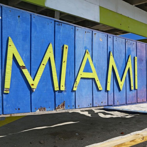 Weathered blue wooden sign spelling "Miami" in yellow letters, set against a sandy area under a building overhang.