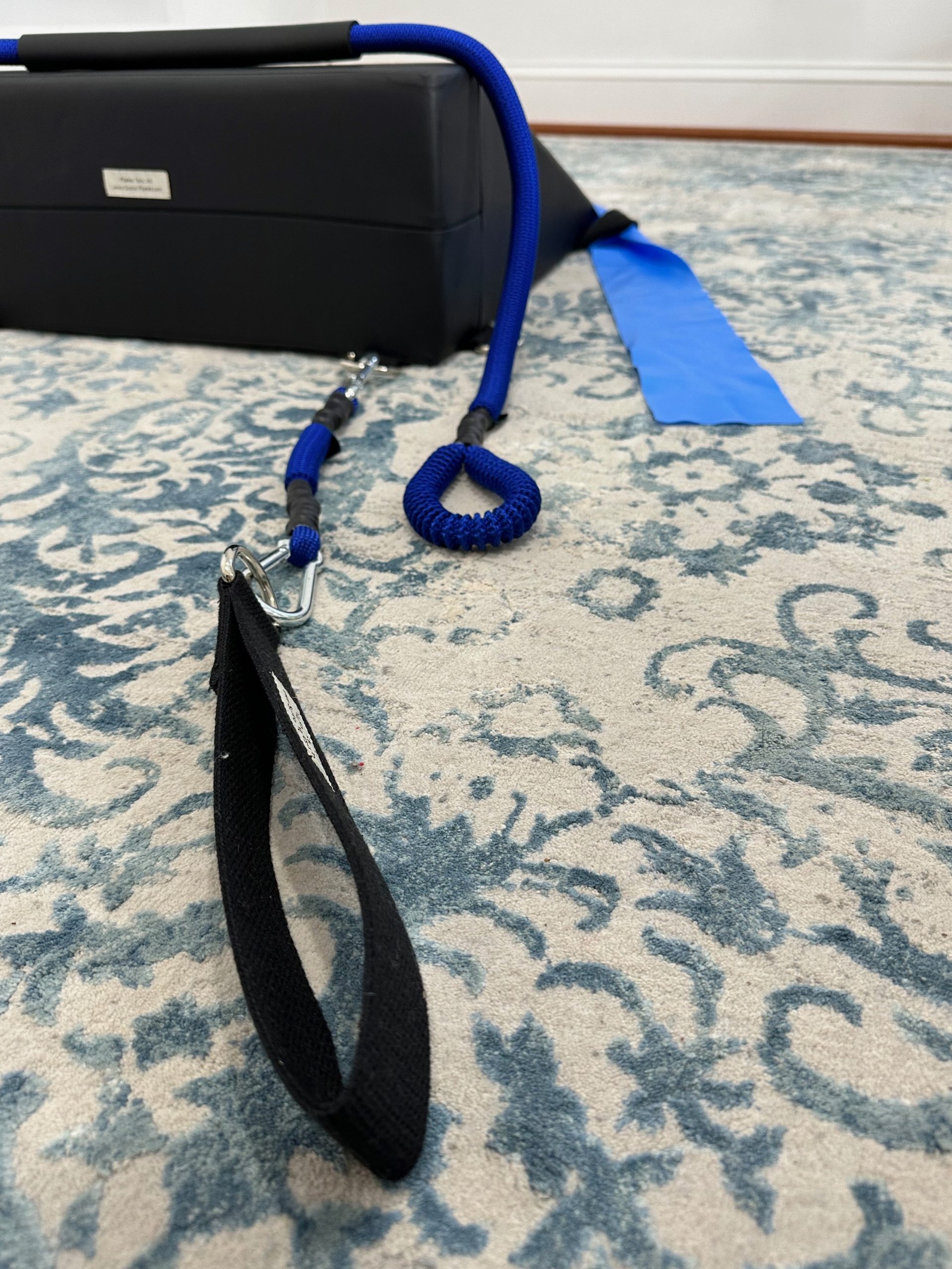 Back view of the K-Fit wedge with the Pilates Teck, Inc. label visible, showing how the resistance cords connect via carabiner clips to fabric handles for exercise.