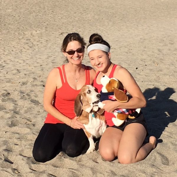 Karena Thek and Olivia kneeling on the beach with Gus, a basset hound, sunny day, everyone smiling.