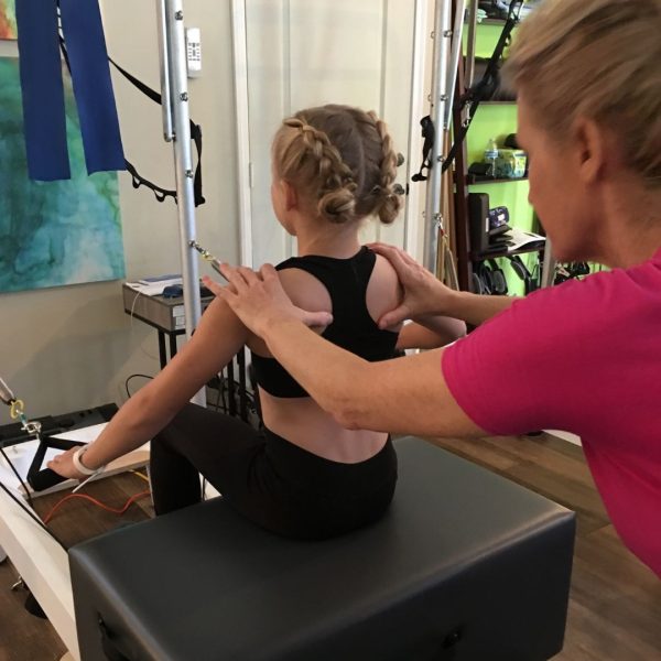 A Scolio-Pilates® practitioner guiding a young girl through a corrective exercise on Pilates equipment, hands on her shoulders.