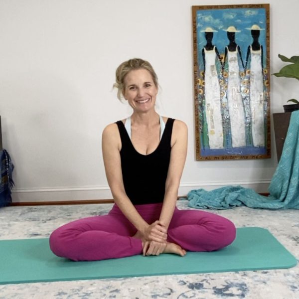 Karena Thek seated cross-legged on a teal mat, smiling, in a warmly decorated room with plants and artwork.