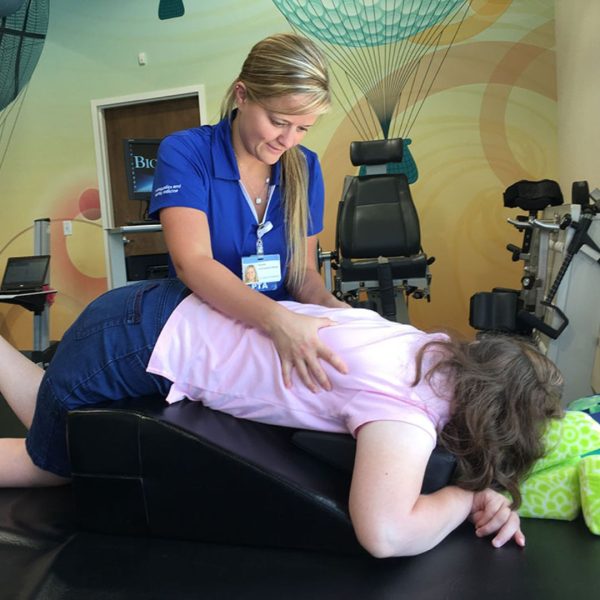 A physical therapist guiding a young girl through a scoliosis-specific exercise over a positioning wedge in a children's hospital setting.