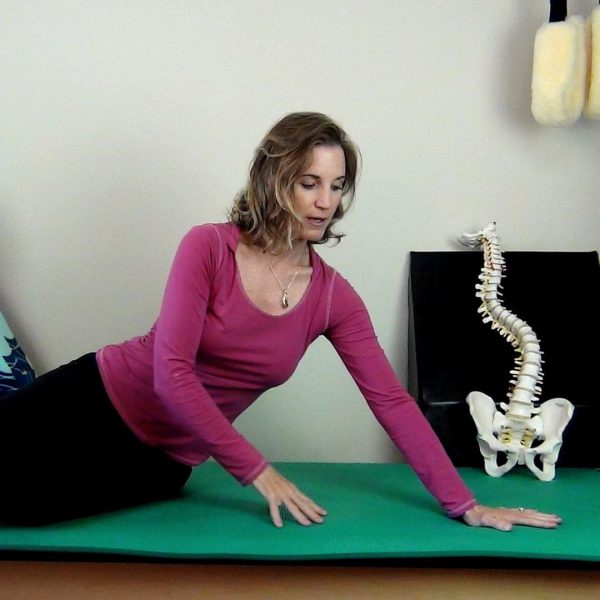 Karena Thek performing a scoliosis-specific side-reaching exercise on a mat, seated beside a model scoliotic spine.