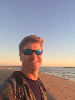 John Herbert, Vice-President of Operations for Scolio-Pilates, smiling outdoors at sunset on the beach
