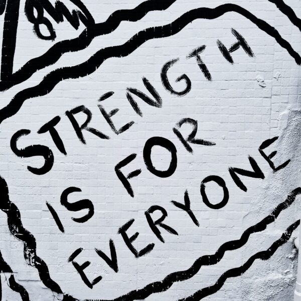 Black and white street mural reading "Strength Is For Everyone" in hand-painted lettering surrounded by a wavy border.