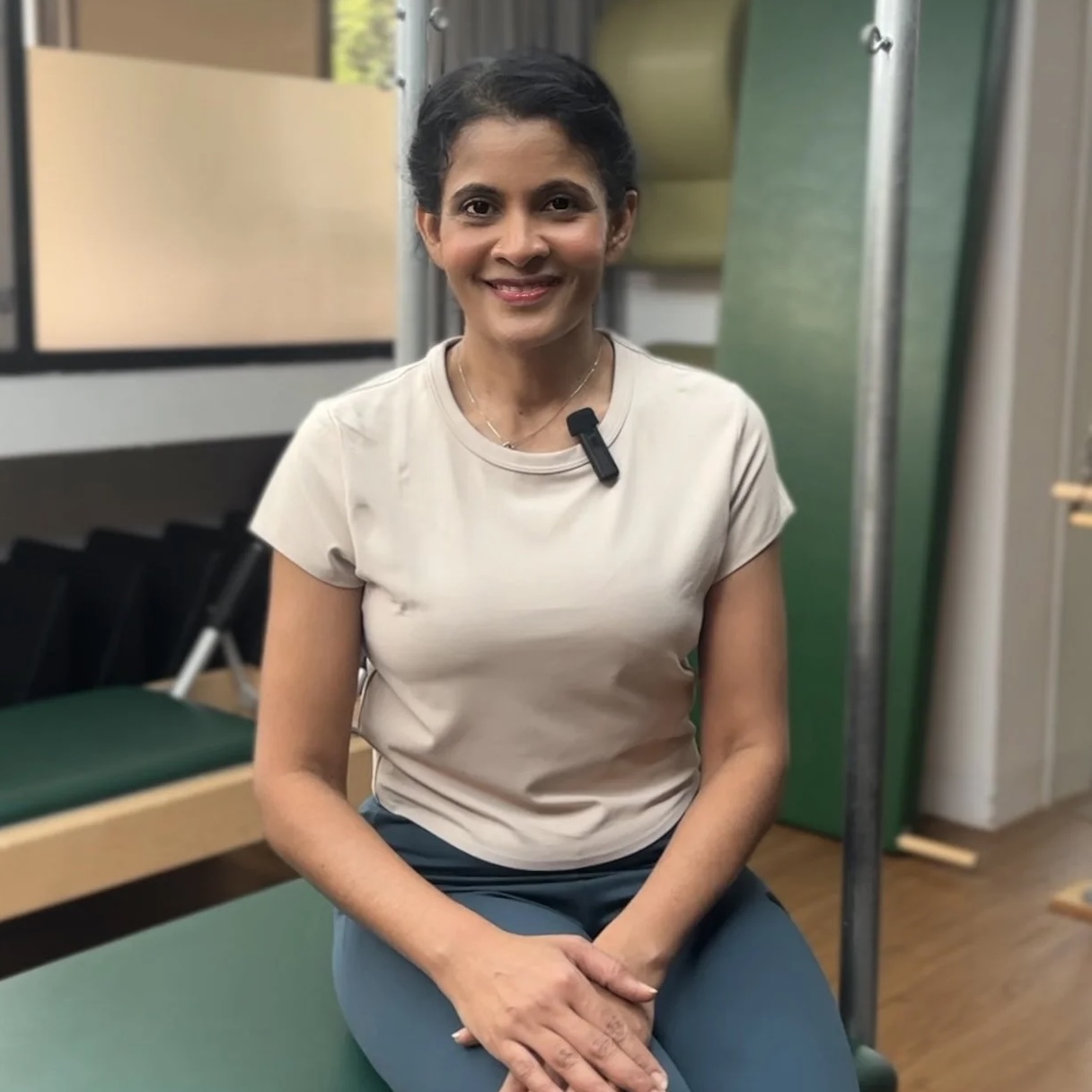 Devahi Palany smiling, seated on Pilates equipment in a studio in Singapore, wearing a microphone.