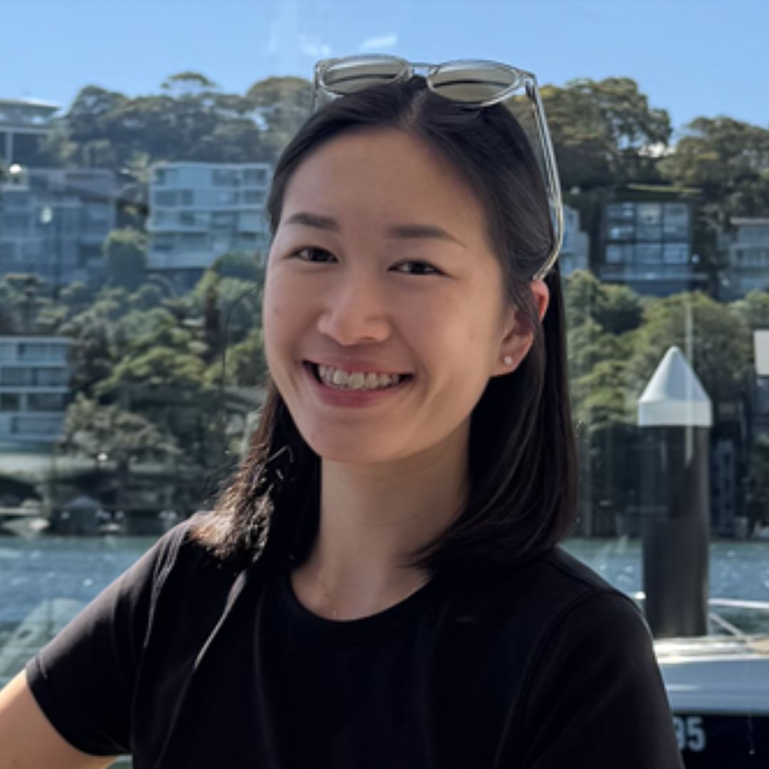Elsie smiling on a boat, waterfront buildings and trees visible in the background, sunglasses resting on her head.