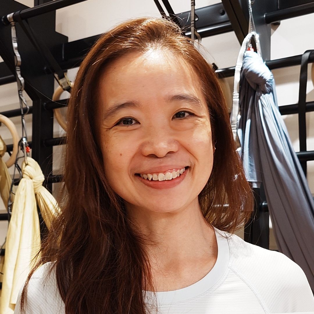 Headshot of Grace Liew, a Scolio-Pilates practitioner, smiling in a Pilates studio with reformer straps and gymnastic rings visible in the background.