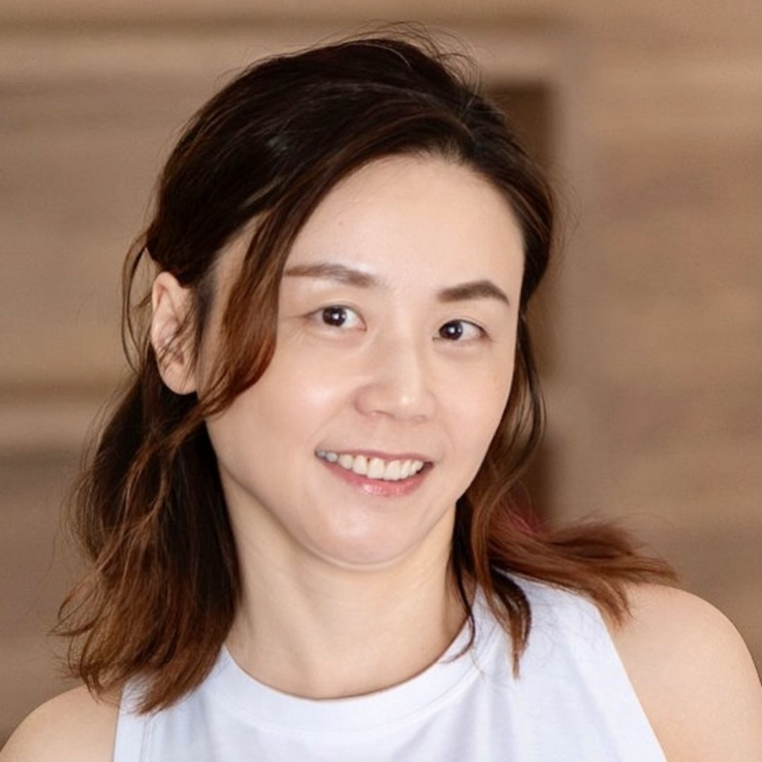 Ivy Pan smiling and looking off to the side, wearing a white tank top, warm wooden background.