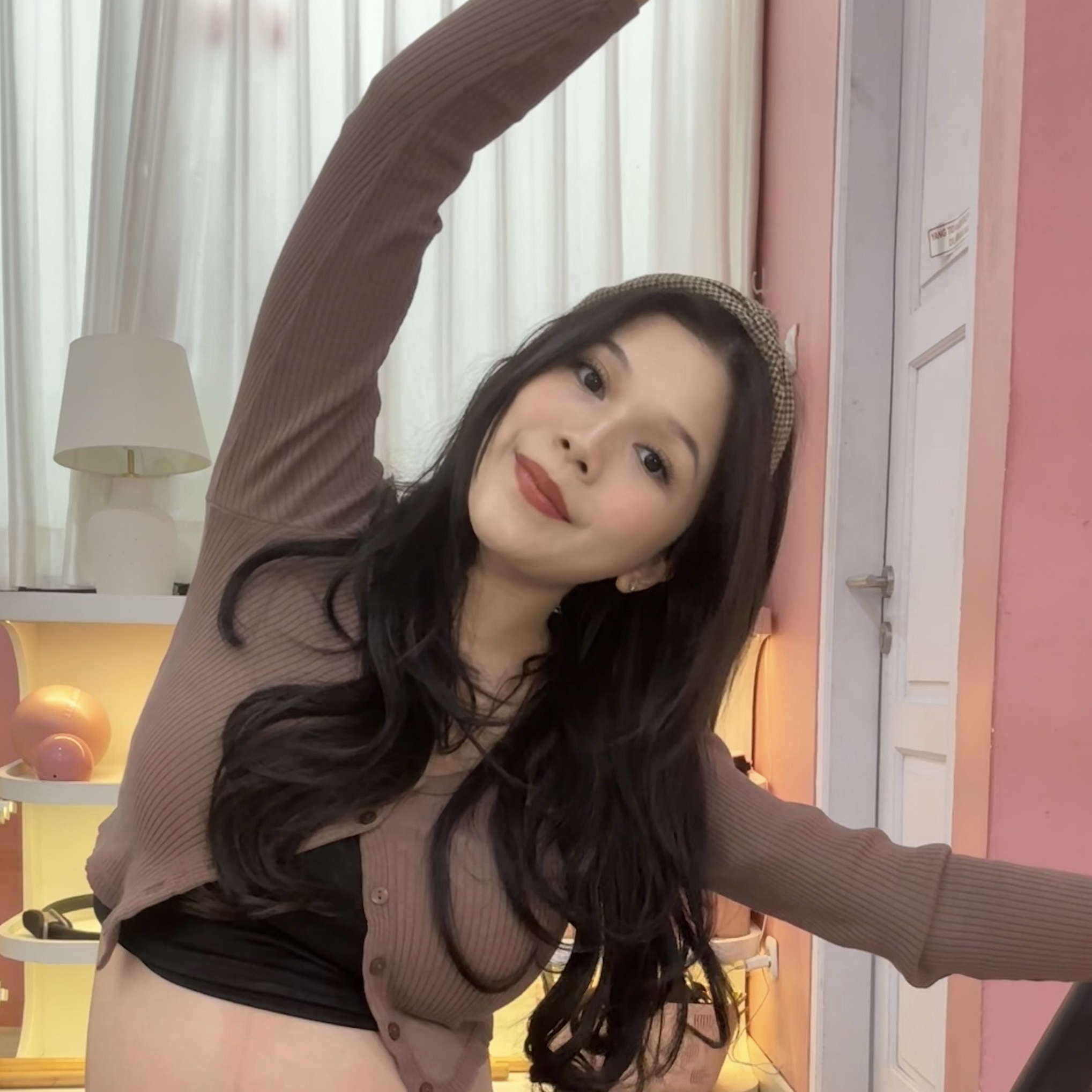 Maya Kartowisastro smiling with one arm raised overhead in a stretch, wearing a brown cardigan, indoors.